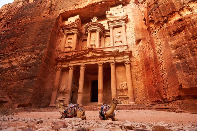 A photo taken from a low angle looking up at the entrance to the Treasury building. The photo shows the pillars and decorations carved into the canon's face. Two camels are seated in front of the entrance.