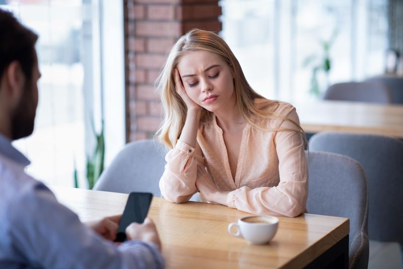 A bored looking woman sits at a cafe table opposite a man on his phone who appears to be ignoring her. 