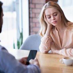 A bored looking woman sits at a cafe table opposite a man on his phone who appears to be ignoring her. 