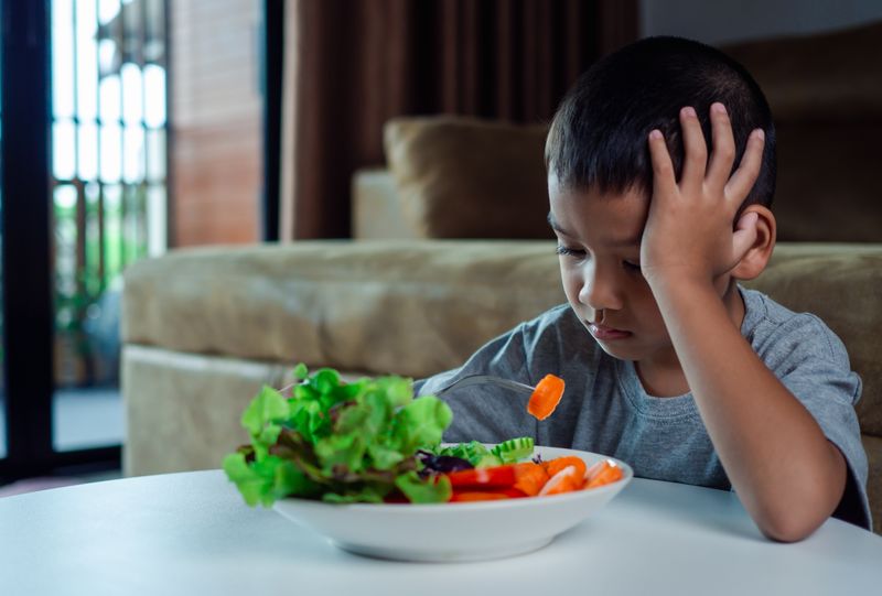 young boy sitting at table looking sad eating a salad