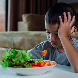 young boy sitting at table looking sad eating a salad