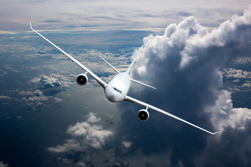 White passenger jet plane in the sky. Aircraft flying high above the cumulus clouds and ocean waves. Airplane front view and right roll.