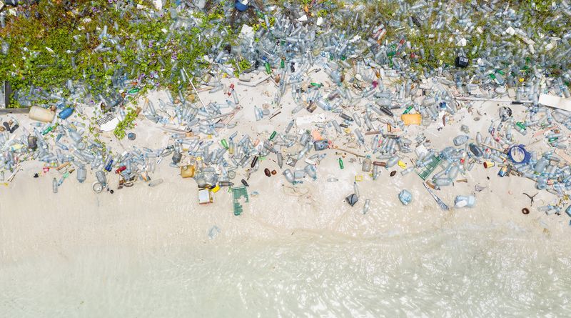 An aerial photo showing masses of plastic bottles washed up on a shoreline. The bottom of the image shows the water's edge and the white sand, but above this line are thousands of bottles and other forms of plastic. 