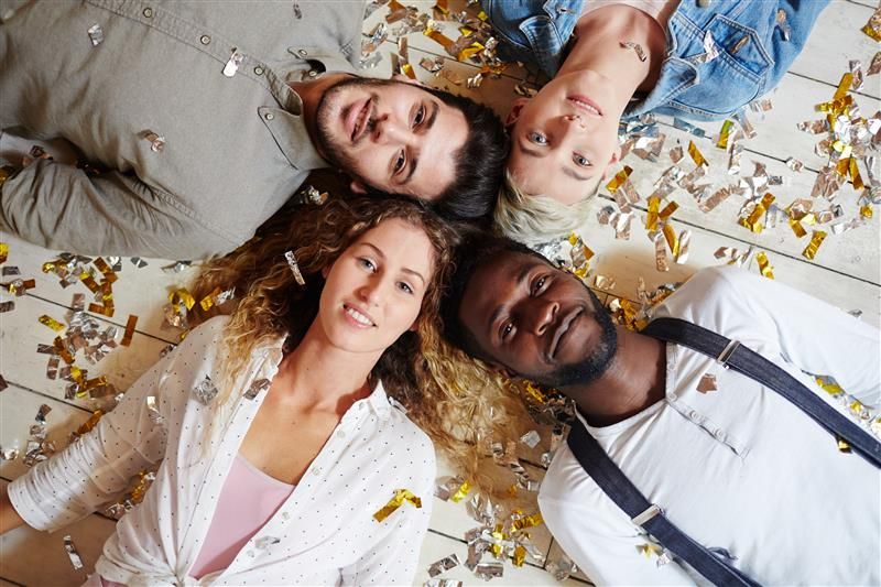 Four adults lay on the floor looking up at the camera. They are arranged with their heads together, their bodies extending off in opposite directions. The first man is white and wearing a grey-ish shirt, his head is next to that of a short-blond haired woman wearing a denim shirt. Next to her is a black man with short hair, a white grandad shirt with braces, and finally another women with long curly hair, wearing a white shirt over a pink t-shirt. They are laying on white floorboards and they are covered in and surrounded by golden confetti. 