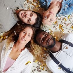 Four adults lay on the floor looking up at the camera. They are arranged with their heads together, their bodies extending off in opposite directions. The first man is white and wearing a grey-ish shirt, his head is next to that of a short-blond haired woman wearing a denim shirt. Next to her is a black man with short hair, a white grandad shirt with braces, and finally another women with long curly hair, wearing a white shirt over a pink t-shirt. They are laying on white floorboards and they are covered in and surrounded by golden confetti. 