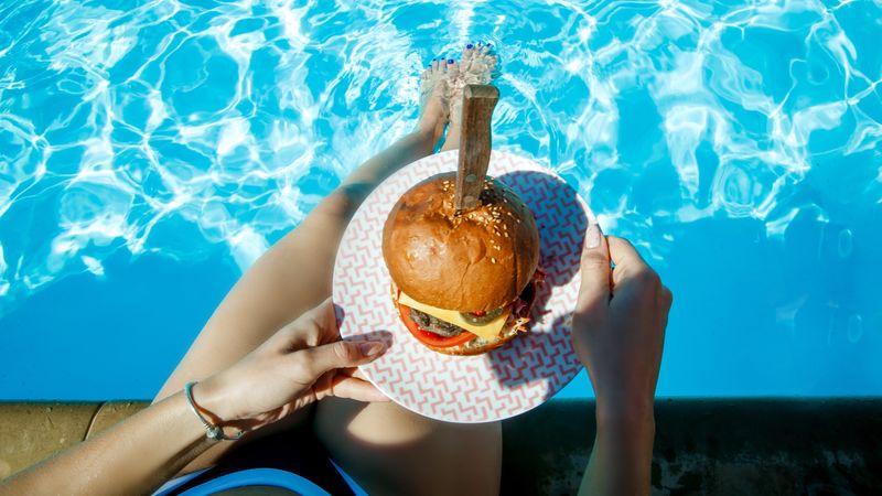 Top view of juicy Burger in girl's hands and feet in the pool. 