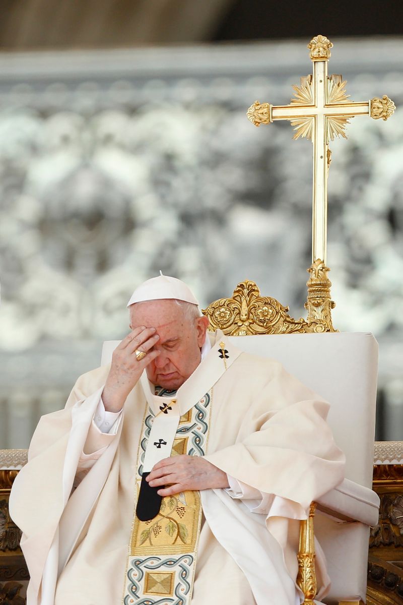 Pope Francis in a white and gold robe sitting in front of a gold cross, touching his forehead