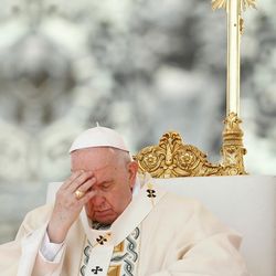 Pope Francis in a white and gold robe sitting in front of a gold cross, touching his forehead