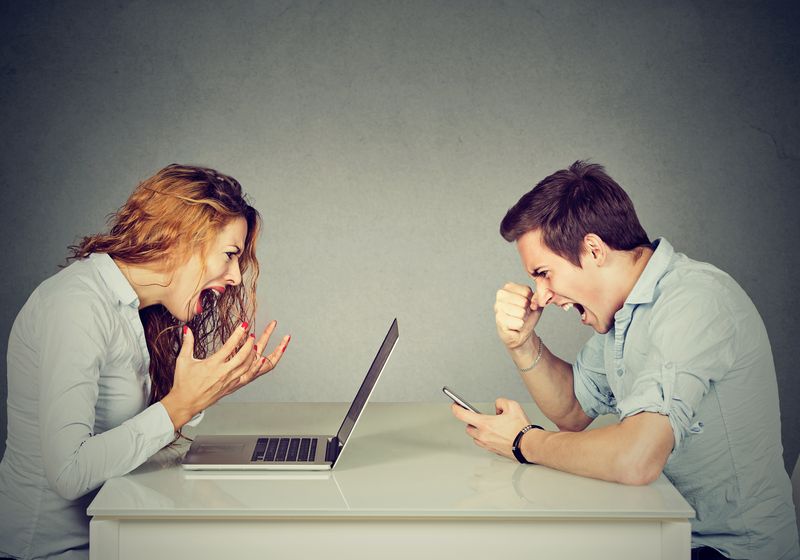 man and woman sitting opposite each other on a table looking angrily at a phone and laptop respectively