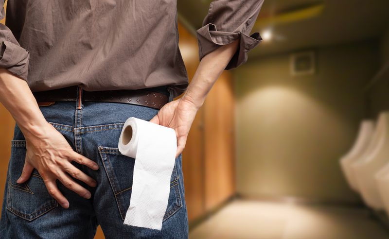 close up of person holding buttocks and a roll of toilet paper; they are photographed with their back to the camera and in the background you can see a blurry bathroom with several urinals and stalls