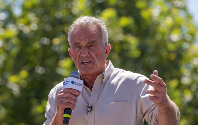 Robert F Kennedy Jr speaking in Des Moines Iowa in August 2023; he is pictured wearing a light beige shirt holding a mic with green trees in the background