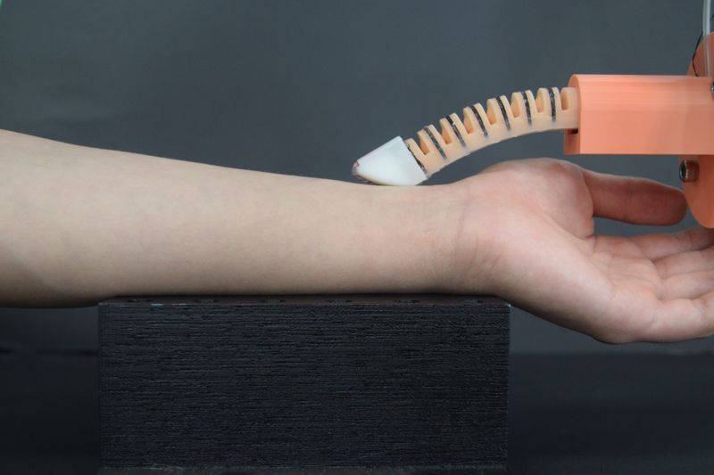 A photo showing the new robotic finger resting on a person's wrist as it takes their pulse. The finger looks curved and make up of segments similar to a spinal cord. At its end is a white tip that is shaped with a flatter surface. The finger itself is connected to an orange plastic structure.