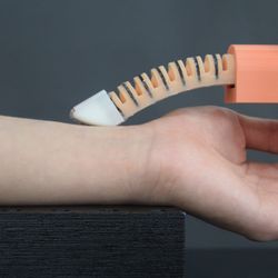 A photo showing the new robotic finger resting on a person's wrist as it takes their pulse. The finger looks curved and make up of segments similar to a spinal cord. At its end is a white tip that is shaped with a flatter surface. The finger itself is connected to an orange plastic structure.