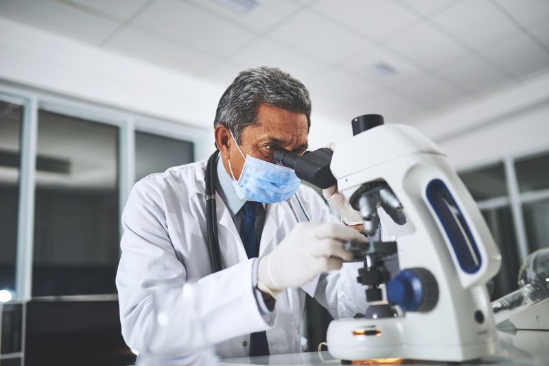 scientist wearing white coat, gloves and surgical mask looking into a microscope