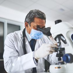 scientist wearing white coat, gloves and surgical mask looking into a microscope