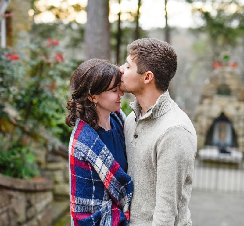 heterosexual couple standing close together outdoors, with green plants and red flowers out of focus in the background near stone steps and a metal gate. She is wearing a blue dress and a red and white plaid shawl, he is wearing a cream sweater and kissing her on the forehead.