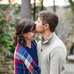 heterosexual couple standing close together outdoors, with green plants and red flowers out of focus in the background near stone steps and a metal gate. She is wearing a blue dress and a red and white plaid shawl, he is wearing a cream sweater and kissing her on the forehead.