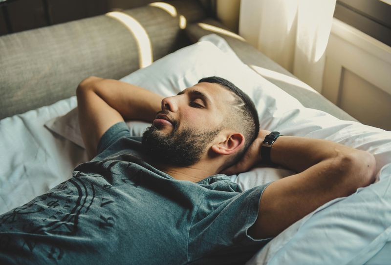 man lying on his back with arms behind his head on a bed, sleeping; he is wearing a green t-shirt and the bed covers are white