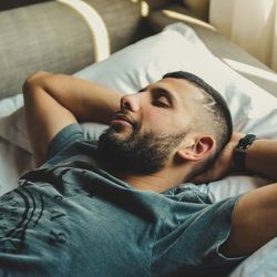 man lying on his back with arms behind his head on a bed, sleeping; he is wearing a green t-shirt and the bed covers are white