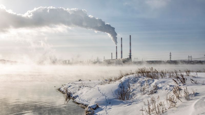 View of Zainskaya TPP with smoking chimneys and an ice-free reservoir with steam rising from the water on a sunny winter frosty day