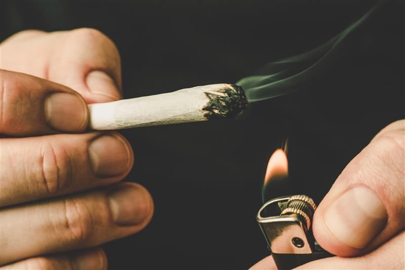 The photo shows a close up of a hand holding a spliff and another holding a lighter flame just below its end. The spliff has been lit and a small length of smoke extends from its end. The background in black. 