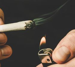 The photo shows a close up of a hand holding a spliff and another holding a lighter flame just below its end. The spliff has been lit and a small length of smoke extends from its end. The background in black. 