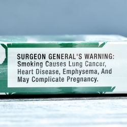 cigarette stubbed out next to packet of cigarettes with surgeon general's warning label about health risks clearly visible