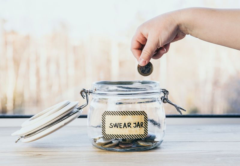 hand placing Euro coin in a glass jar labelled "Swear Jar"