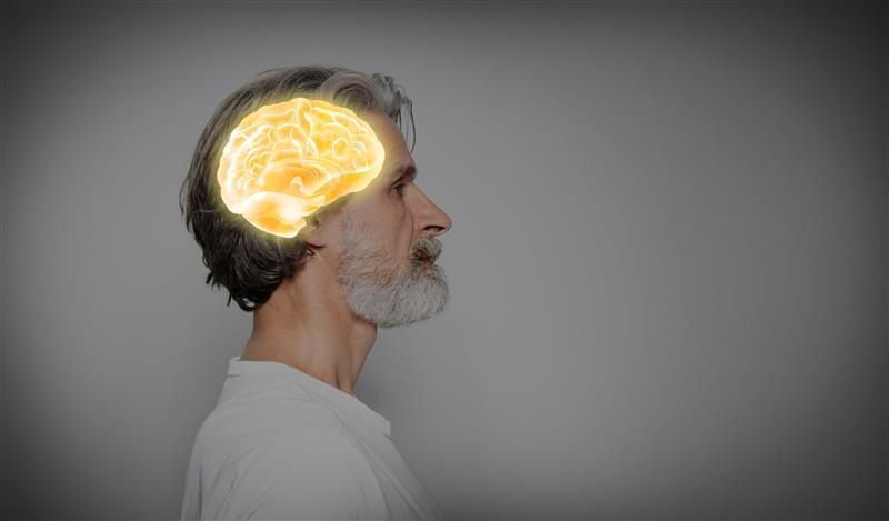 The photo shows an older man with a short white beard and wearing a white narrow-collared shirt standing on profile, facing the right. The man has a brain image superimposed on his head, the brain is a yellow-golden colour with white that highlights its shape and details.  