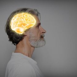 The photo shows an older man with a short white beard and wearing a white narrow-collared shirt standing on profile, facing the right. The man has a brain image superimposed on his head, the brain is a yellow-golden colour with white that highlights its shape and details.  