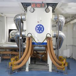 A photo showing the resistive magnet. It looks like a cylindrical body on four tubular legs. It has two sets of brown cables running from its bottom, two on each side, and two thick silver tubes on each side positioned in the middle and at the top of the device. The whole thing is located reaches from floor to ceiling in a room with a blue floor.