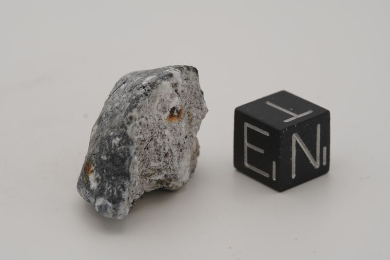  A rare aubrite meteorite: a greyish rock with a few spots of brown is visible next to a little black cube