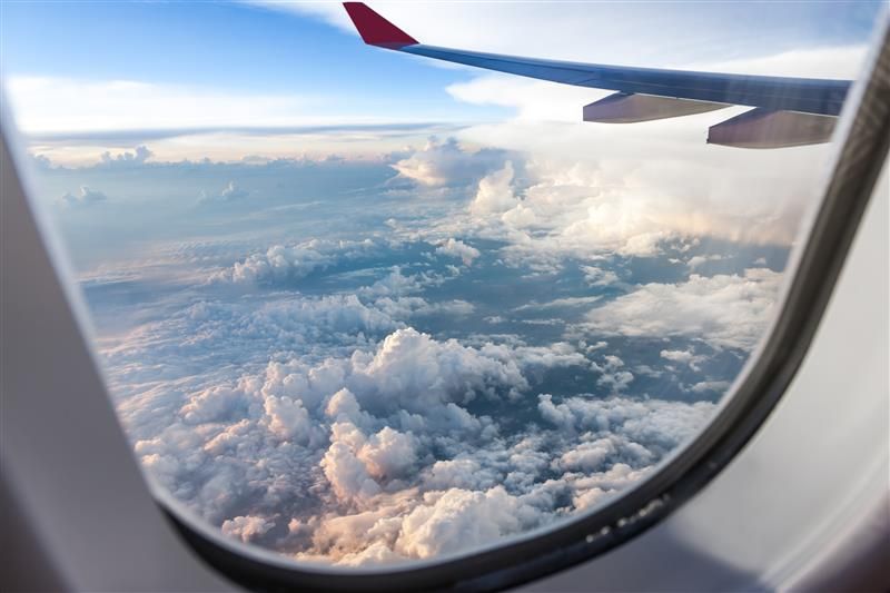 An photo take from a plane window. The shot shows an accumulation of clouds below the plane that look like bubbly mountains. The plane's left wing is visible in the top right of the photo.  