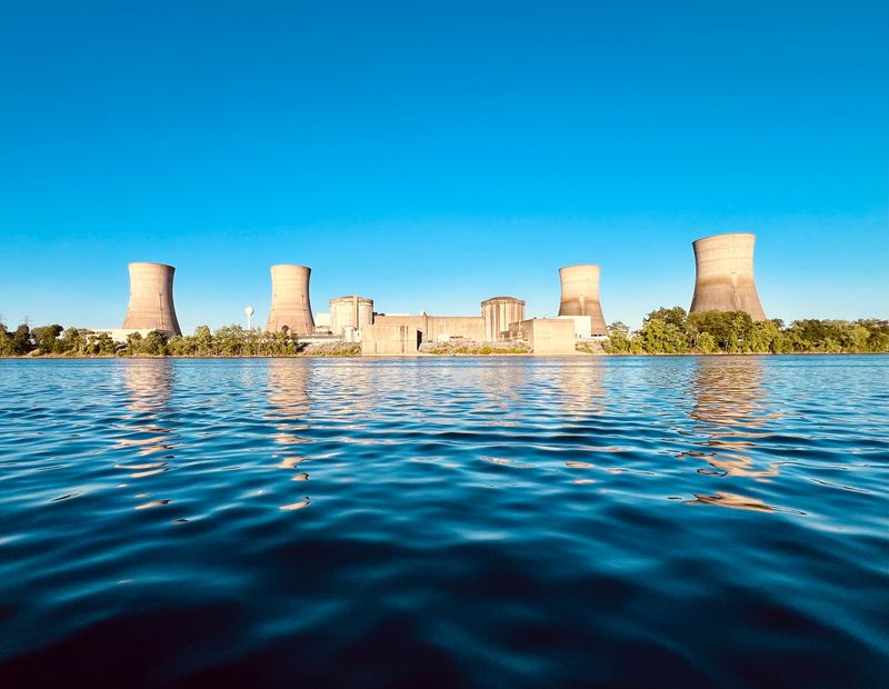 A photo take from across the water showing the Three Mile Island power plant. The plant's four cooling towers are visible as they are clustered around the main reactor buildings. The photo was taken on a bright day and the sky behind the plant is completely blue.