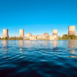 A photo take from across the water showing the Three Mile Island power plant. The plant's four cooling towers are visible as they are clustered around the main reactor buildings. The photo was taken on a bright day and the sky behind the plant is completely blue.