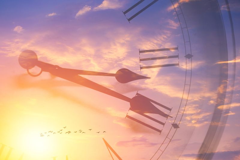 sunset sky with distant flock of birds, overlaid with image of analog clock to illustrate the passage of time