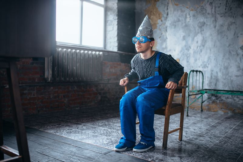 man wearing a tinfoil dunce's cap and hypnotising glasses, leaning forward in a chair in an empty room, wearing blue dungarees and a grey sweater