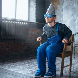 man wearing a tinfoil dunce's cap and hypnotising glasses, leaning forward in a chair in an empty room, wearing blue dungarees and a grey sweater