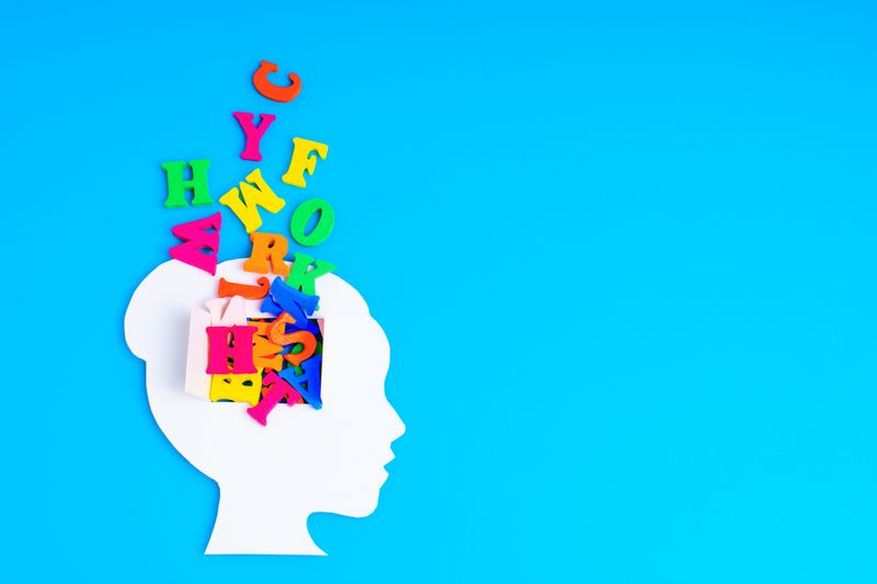 white paper silhouette of human head facing to the right, with a window cut open where the brain would be and multicoloured letters of the English alphabet spilling out onto a bright turquoise background