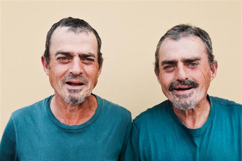 Two older twin men stand next to each other and smile at the camera. They are wearing identical green T-shirts and they have the same short hair and trimmed beards. 