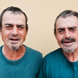 Two older twin men stand next to each other and smile at the camera. They are wearing identical green T-shirts and they have the same short hair and trimmed beards. 