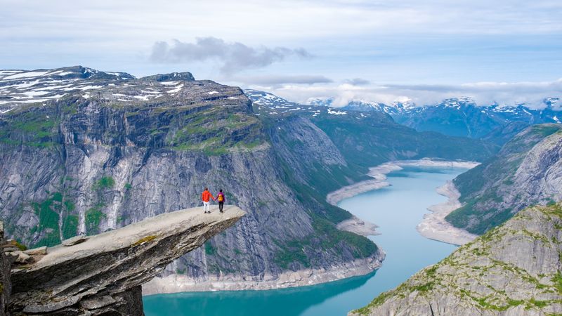 Two hikers stand on the edge of Trolltunga, Norway, a massive cliff overlooking a stunning fjord in Norway.