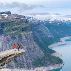 Two hikers stand on the edge of Trolltunga, Norway, a massive cliff overlooking a stunning fjord in Norway.