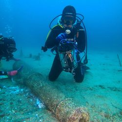 A diver rests on the sea floor in front of the remains of a 16th century cannon. The diver is holding a tubular instrument. Another diver can be seen cleaning end of the cannon in the background. The water visibility is murky and the scene has been lit by an artificial light. 