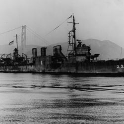 A black and white photo showing USS Stewart in the water with the Golden Gate bridge in the background. The water is calm and the vessel had two crew members standing on its bow. 