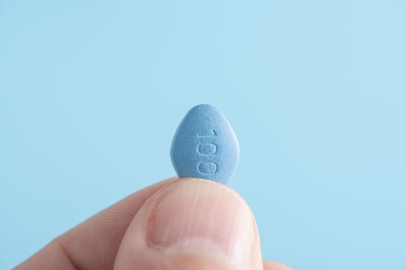 close-up of a person holding a blue viagra pill between their thumb and forefinger on blue background.