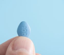 close-up of a person holding a blue viagra pill between their thumb and forefinger on blue background.