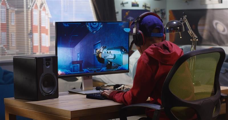 A medium shot of the back of a boy in a red hoodie and blue cap sitting at a desk playing a video game. The boy's room contains posters of planets on the far right of the photo. In the background, you can see a window view of a houses across the road. The video game shows a first person perspective of a someone holding a gun.  