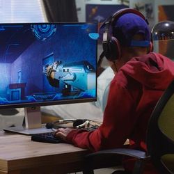 A medium shot of the back of a boy in a red hoodie and blue cap sitting at a desk playing a video game. The boy's room contains posters of planets on the far right of the photo. In the background, you can see a window view of a houses across the road. The video game shows a first person perspective of a someone holding a gun.  