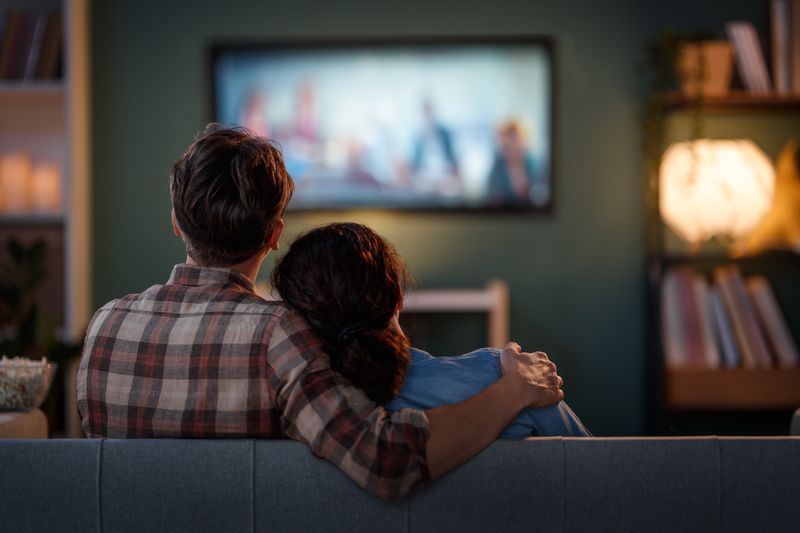 couple viewed from behind as they sit together on the sofa and watch a movie on a wall-mounted TV